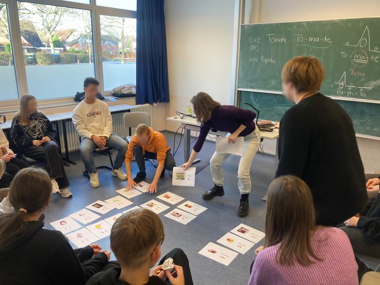 Studierende und Schüler:innen sitzen im Stuhlkreis, während drei Personen aktiv Bildkarten am Boden ordnen. Im Hintergrund ist eine Tafelanschrift zu sehen.