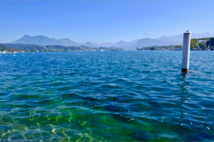 Foto von dem blauen Meer an einem sonnigen Tag. Berge sind im Hintergrund zu sehen und die Sonne spiegelt sich im Wasser. 