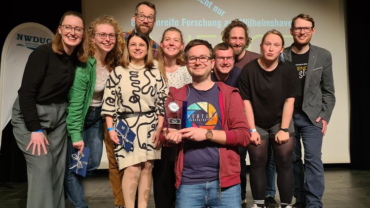 Freuen sich mit Dr. Markus Prinz über den Gewinn des Science Slam: Nathalie Kürten (IfV), Komoderatorin des Abends; Dr. Maren Bertheau (UOL); Dr. Rufina Fingerhut; Dr. Matthias Weissensteiner (IfV); Jenny Neuhaus (DZMB, Senckenberg am Meer); der Preisträger (ICBM); Dominik Antoni (AWI); Lennart Barke (Wattenmeer Besucherzentrum, hielt einen Vortrag über Wissenschaft außer Konkurrenz); Justine Bertram (IfV) und Dr. Moritz Mennenga (NIhK, Geschäftsführer NWDUG).