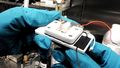 Two hands in blue protective gloves are holding the measuring cell. It is white, about the size of a telephone and connected with cables. 