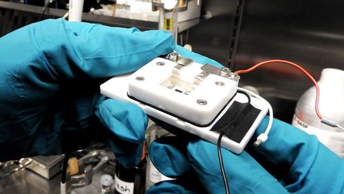 Zwei Hände in blauen Schutzhandschuhen halten die Messzelle. Sie ist weiß, etwa so groß wie ein Telefon und mit einigen Kabeln verbunden. 