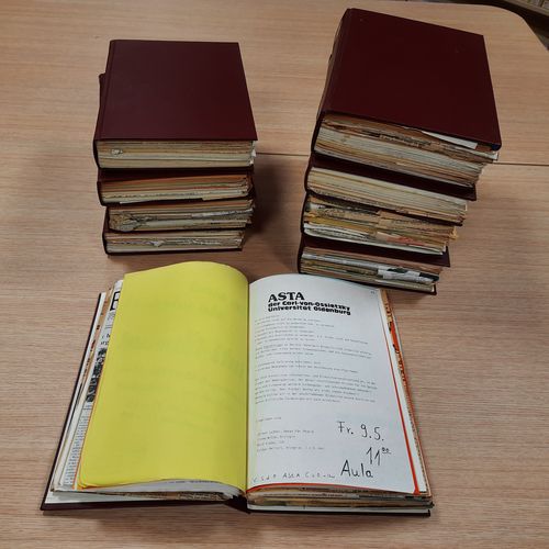 Das Bild zeigt mehrere Bände mit Flugblättern, die auf einem Tisch liegen. Eines der Bücher ist aufgeschlagen. Zu erkennen ist ein Flugblatt des AStA, der zu einer Veranstaltung einlädt. Manche der Flugblätter sind auf weißem, andere auf buntem Papier gedruckt.