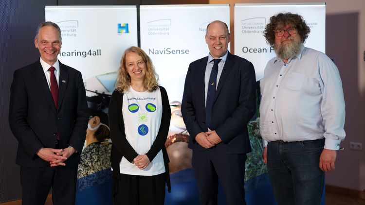 Vier Personen stehen nebeneinander vor drei Roll-ups der einzelnen Cluster. 