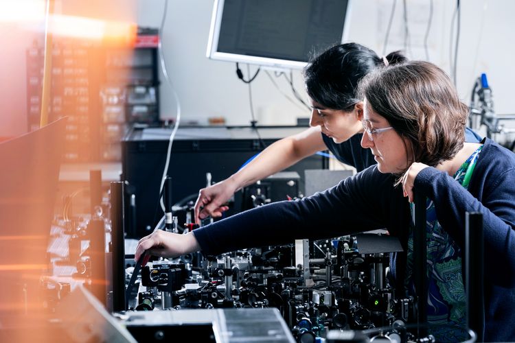 Two women are preparing an experiment in a physics laboratory, setting up microscope-like devices.