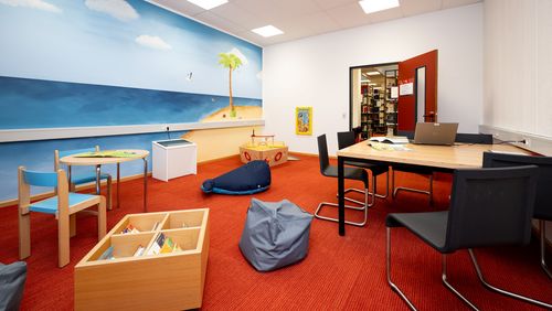 Ein Raum, durch dessen Tür man die Bibliothek im Hintergrund sieht. Ein fahrbares Bücherregal,Sitzsäcke, ein Kindertisch und ein Spielboot bieten Kindern Spielgelegenheiten. Eltern können am Tisch daneben arbeiten. 