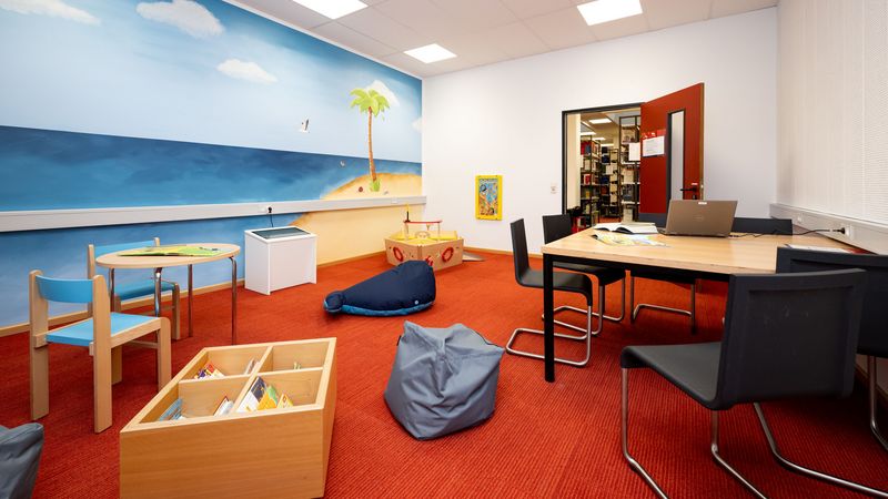 Weitwinkelaufnahme eines Raumes, an dessen Ende eine offene Tür den Blick auf Bücherregale eröffnet. Im Raum findet sich an der rechten Wand ein Tisch mit mehreren Stühlen. Die linke Wand ist bemalt mit einem Meeresmotiv. davor findet sich ein großes Holzsschiff, ein Kindertisch mit zwei Stühlen sowie eine Kiste mit Büchern. Im Raum liegen außerdem ein grauer Sitzsack und ein großer blauer Stoff-Blauwahl. Der Boden ist mit rotem Teppich ausgelegt.