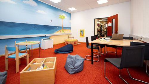 Weitwinkelaufnahme eines Raumes, an dessen Ende eine offene Tür den Blick auf Bücherregale eröffnet. Im Raum findet sich an der rechten Wand ein Tisch mit mehreren Stühlen. Die linke Wand ist bemalt mit einem Meeresmotiv. davor findet sich ein großes Holzsschiff, ein Kindertisch mit zwei Stühlen sowie eine Kiste mit Büchern. Im Raum liegen außerdem ein grauer Sitzsack und ein großer blauer Stoff-Blauwahl. Der Boden ist mit rotem Teppich ausgelegt.