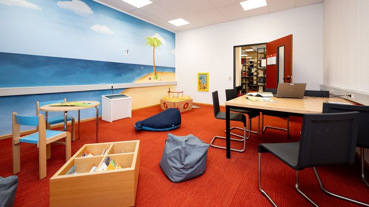 Weitwinkelaufnahme eines Raumes, an dessen Ende eine offene Tür den Blick auf Bücherregale eröffnet. Im Raum findet sich an der rechten Wand ein Tisch mit mehreren Stühlen. Die linke Wand ist bemalt mit einem Meeresmotiv. davor findet sich ein großes Holzsschiff, ein Kindertisch mit zwei Stühlen sowie eine Kiste mit Büchern. Im Raum liegen außerdem ein grauer Sitzsack und ein großer blauer Stoff-Blauwahl. Der Boden ist mit rotem Teppich ausgelegt.