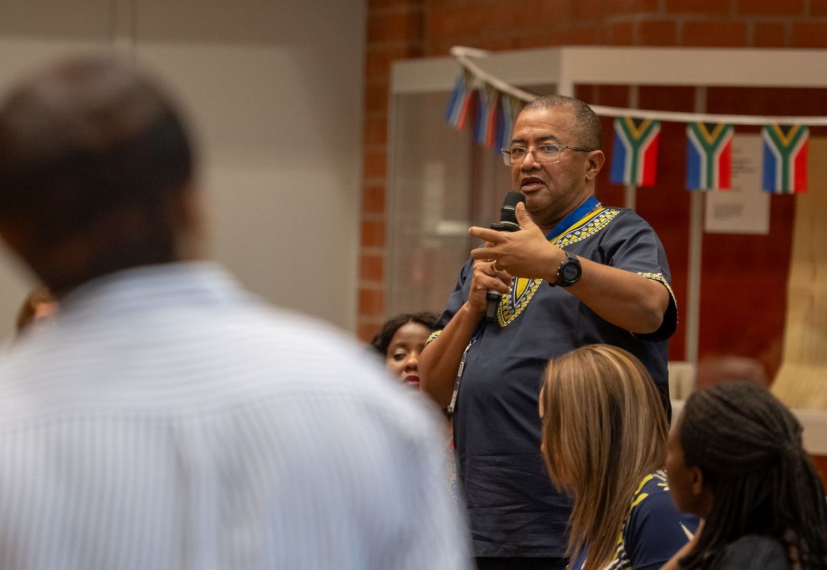 Symposium Ein Mann mit Brille und einem blauen Oberteil mit gelben Mustern hält ein Mikrofon und spricht. Er blickt in Richtung des Betrachters und gestikuliert mit seiner rechten Hand. Im Hintergrund sind weitere Personen und eine Girlande mit den Farben der südafrikanischen Flagge zu sehen. Das Bild vermittelt den Eindruck einer Rede oder einer Frage-und-Antwort-Runde während des Symposiums. Der Mann scheint eine wichtige Rolle bei der Veranstaltung zu spielen.