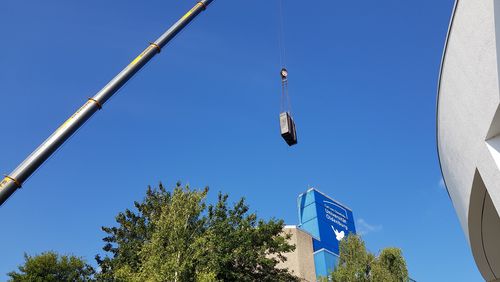 Blick in den Himmel, im Vordergrund hängt ein Rechnerschrank am Kran, im Hintergrund Bäume und das Gebäude A1 mit Friedenstaube, rechts ragt die runde Wand des Hörsaalzentrums noch ins Bild. 