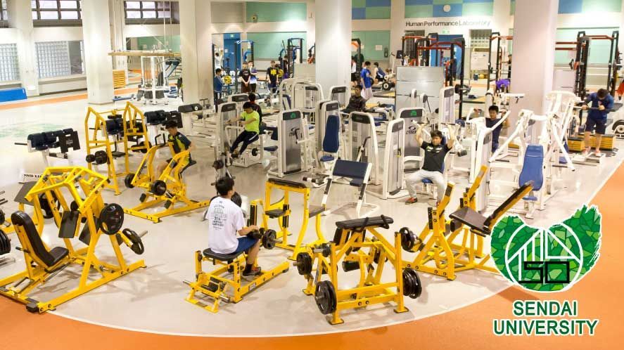 Das Bild zeigt einen geräumigen Fitnessraum, der mit einer Vielzahl von Trainingsgeräten ausgestattet ist. Mehrere Personen sind dabei zu sehen, wie sie verschiedene Übungen an den Geräten durchführen, darunter Beinpresse, Rudermaschinen und Kraftstationen. Der Raum ist hell erleuchtet und wirkt gut ausgestattet, was auf ein umfassendes Angebot für sportliche Aktivitäten hindeutet.