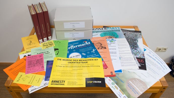 Auf einem Tisch lieben mehrere bunte Flugblätter, Plakate und Broschüren übereinander, hinten stehen drei Bände und zwei Kartons der Flugblattsammlung. 