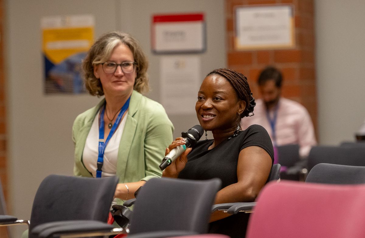 Symposium Eine Frau mit geflochtenem Haar hält ein Mikrofon und lächelt in eine bestimmte Richtung. Neben ihr sitzt eine weitere Frau mit Brille und einem hellgrünen Jackett. Im Hintergrund ist eine weitere Person und Poster an einer roten Backsteinwand zu sehen. Das Bild zeigt eine entspannte Atmosphäre während des Symposiums, möglicherweise während einer Fragerunde oder einer Diskussion. Die Frau mit dem Mikrofon scheint aktiv an der Veranstaltung teilzunehmen.