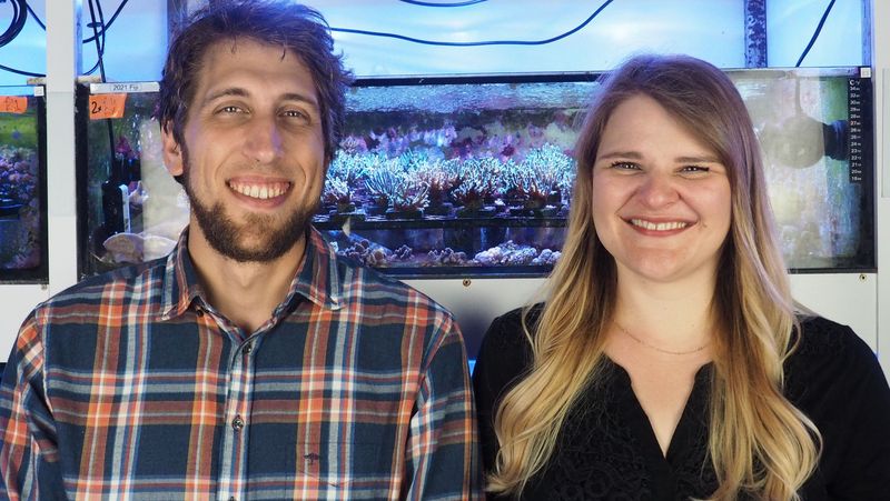 Das Bild zeigt Samuel Nietzer und Mareen Möller. Sie stehen vor einem Aquarium, in dem sich Korallen befinden.