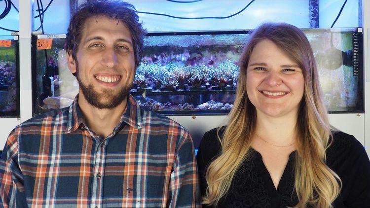 Das Bild zeigt Samuel Nietzer und Mareen Möller. Sie stehen vor einem Aquarium, in dem sich Korallen befinden.