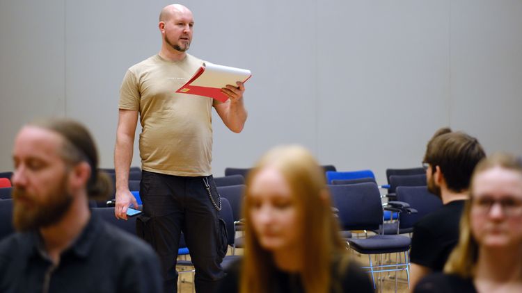 Das Bild zeigt Kai Janssen. Er steht vor einer Stuhlreihe im BIS-Saal und hält eine Mappe mit Text vor sich. Daraus liest er vor. Im Vordergrund sind einige Darstellerinnen und Darsteller zu erkennen. Die meisten von ihnen sitzen mit dem Rücken zu Janssen.