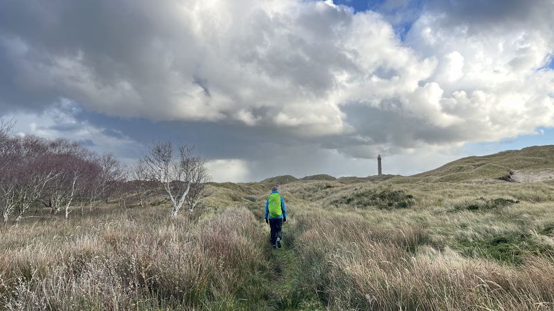 Person mit Rucksack ist von hinten zu sehen, wie sie einen schmalen Pfad durch die karge Dünenlandschaft läuft. Am Horizont ist der Leuchtturm zu sehen. 