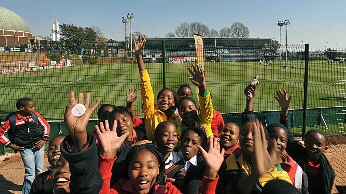 Das Bild zeigt eine Gruppe fröhlicher Kinder, die vor einem Fußballstadion stehen und in die Kamera lächeln und winken. Im Hintergrund ist ein grünes Fußballfeld mit Zuschauerrängen und Werbebannern zu sehen, und im weiteren Hintergrund erheben sich Gebäude und Lichtmasten
