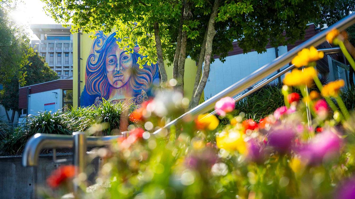 Das Bild zeigt ein farbenfrohes Wandgemälde einer Frau mit langen, gewellten Haaren, das an der Seite eines Gebäudes auf dem Campus der University of Waikato gemalt ist. Im Vordergrund sind bunte Blumen und eine Metallgeländerstange zu sehen, die den Blick auf das Kunstwerk lenken und eine lebendige Atmosphäre schaffen.