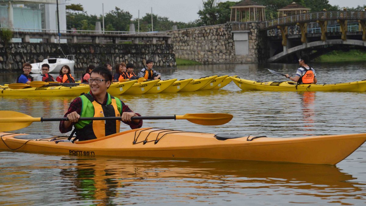 Das Bild zeigt eine Gruppe von Studierenden, die auf einem See oder Fluss Kajak fahren. Im Vordergrund ist ein lächelnder Studierender in einem gelben Kajak zu sehen, während im Hintergrund weitere Kajaks mit anderen Studierenden zu erkennen sind. Alle tragen Schwimmwesten. Die Szene vermittelt eine fröhliche und aktive Atmosphäre und zeigt eine Freizeitaktivität, die von der Universität angeboten wird.
