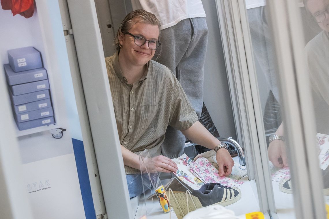 Das Bild zeigt Edwin Mårtensson. Er sitzt auf einer Auslage hinter einem Schaufenster und ist von diversen Textilien, Schuhen und anderen Gegenständen umgeben. Er lächelt in die Kamera.