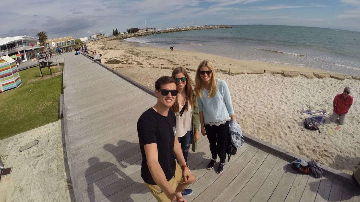 Das Bild zeigt drei lächelnde Personen, die auf einem Holzsteg am Strand posieren. Im Hintergrund erstreckt sich ein Sandstrand mit Wellen und Menschen, die am Strand spazieren gehen. Der Himmel ist leicht bewölkt und die Sonne scheint. Die Szene wirkt entspannt und fröhlich.