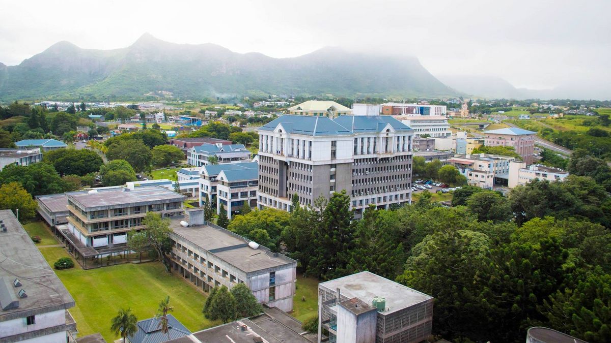  Eine Vogelperspektive zeigt den Campus der University of Mauritius vor dem Hintergrund grüner Berge. Mehrere Gebäude der Universität sind zwischen Bäumen und Grünflächen zu sehen. Im Hintergrund erstreckt sich eine Stadtlandschaft.