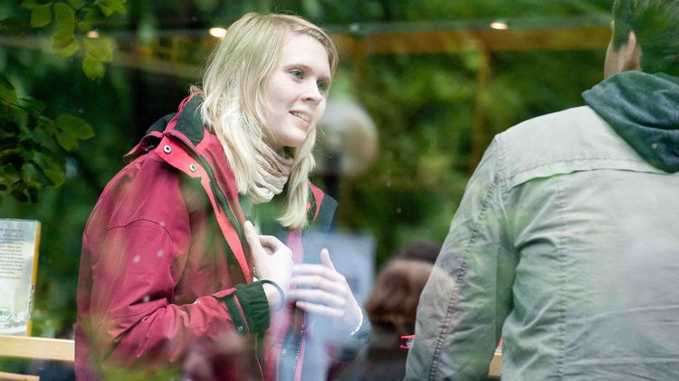 Cafe-Bar Campus Haarentor Eine Frau und ein Mann werden durch eine Glasscheibe betrachtet. Die Frau mit roter Jacke gestikuliert.