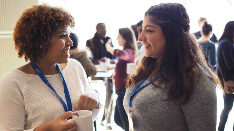 Studierende Zwei junge Frauen unterhalten sich in einem hellen Raum und tragen Namensschilder.