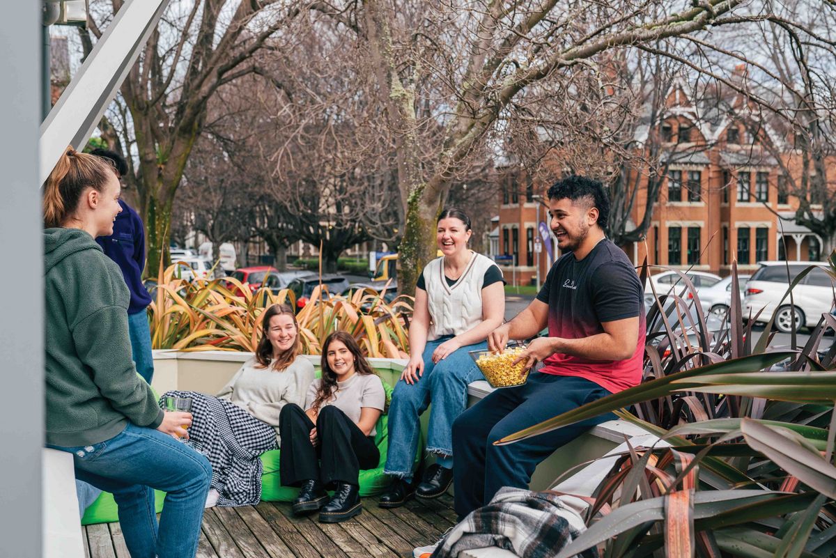 Das Bild zeigt eine Gruppe von Studierenden, die entspannt auf einer Holzterrasse im Freien sitzen und sich unterhalten. Sie lachen und genießen Snacks, was eine freundliche und einladende Atmosphäre vermittelt. Im Hintergrund sind Bäume und Gebäude zu sehen, die darauf hindeuten, dass die Szene auf dem Campus der University of Otago spielt.
