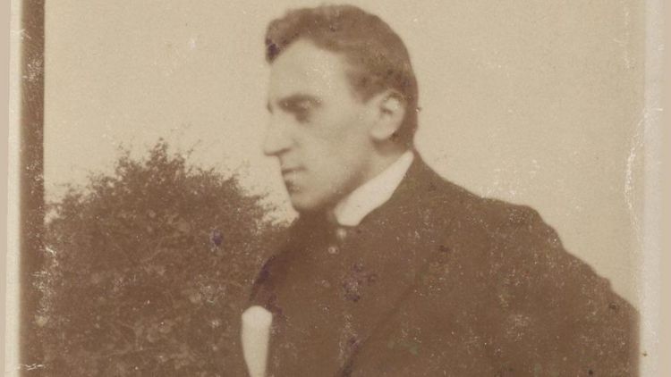 Das sepiafarbene Bild zeigt Ossietzky in seinen jungen Jahren. Er trägt einen Anzug und hat die Hände in die Hüften gestemmt. Er schaut zur Seite.
