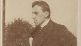 Das sepiafarbene Bild zeigt Ossietzky in seinen jungen Jahren. Er trägt einen Anzug und hat die Hände in die Hüften gestemmt. Er schaut zur Seite.
