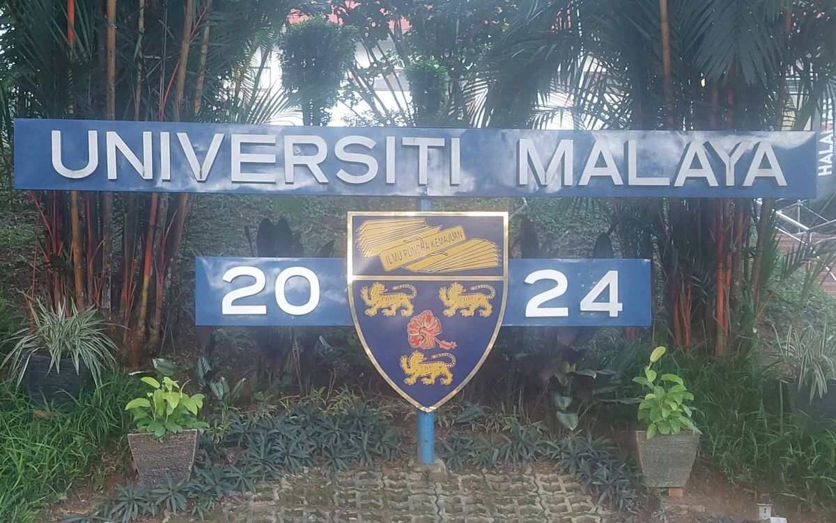 Das Bild zeigt ein Schild mit dem Schriftzug "Universiti Malaya" (Universität Malaya) vor üppiger grüner Vegetation. Das Schild ist blau und enthält das Wappen der Universität sowie die Jahreszahlen 2024. Im Hintergrund sind Bäume und Gebäude zu sehen. Die Szene vermittelt einen Eindruck von akademischer Tradition und einer grünen Campusumgebung.