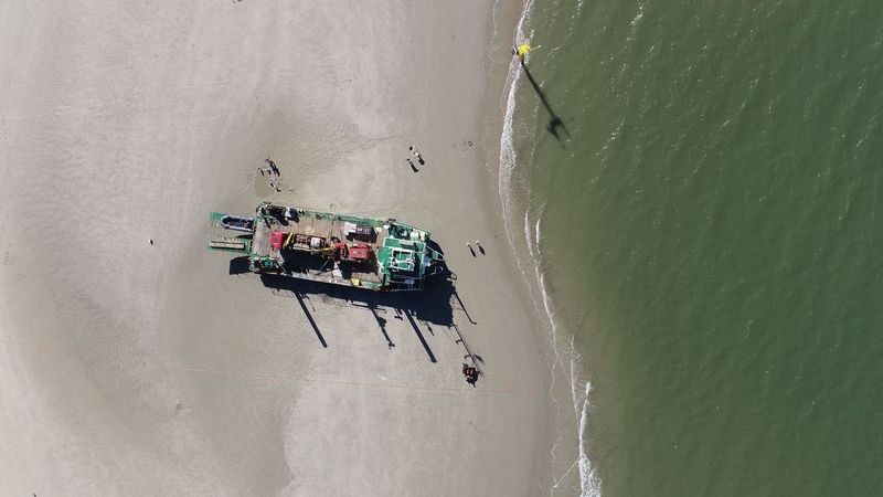 Das Bild zeigt eine Drohnenaufnahme eines Schiffs mit Bohrausstattung, das auf dem Strand von Spiekeroog trocken auf dem Sand liegt. Auf der rechten Seite sind die Nordsee und ein gelber Messpfahl der Oldenburger Forschungsgruppe DynaDeep zu erkennen.