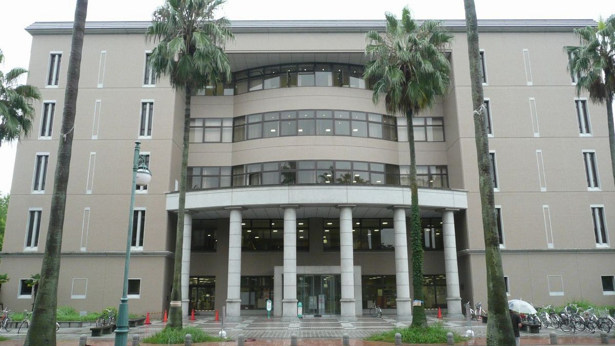 Das Bild zeigt die Fassade eines beigefarbenen Gebäudes der Kagoshima Universität mit einer geschwungenen, offenen Eingangshalle, die von Säulen getragen wird. Mehrere Palmenbäume stehen vor dem Gebäude und rahmen den Eingangsbereich ein. Vor dem Gebäude befinden sich Fahrradständer mit einigen abgestellten Fahrrädern und ein paar orangen Verkehrskegeln.