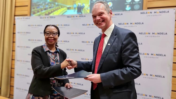 Die Vizekanzlerin der Nelson Mandela University (NMU), Prof. Dr. Sibongile Muthwa, und der Präsident der Universität Oldenburg, Prof. Dr. Ralph Bruder, reichen einander mit Blick in die Kamera die Hand. In ihrer jeweils linken Hand halten sie die neue Kooperationsvereinbarung. Im Hintergrund ist ein Aufsteller mit dem Schriftzg und Logo der NMU.