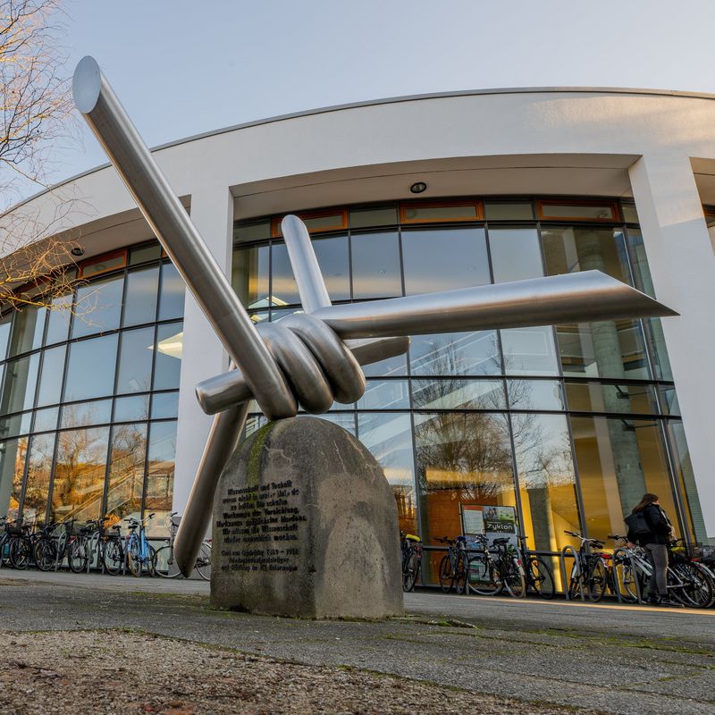 Das Bild zeigt das Mahnmal für Ossietzky vor dem Audimax. Es stellt einen Stacheldraht dar. Vorne zu erkennen ist die im Text erwähnte Inschrift. Im Hintergrund stehen Fahrräder, die Sonne scheint.