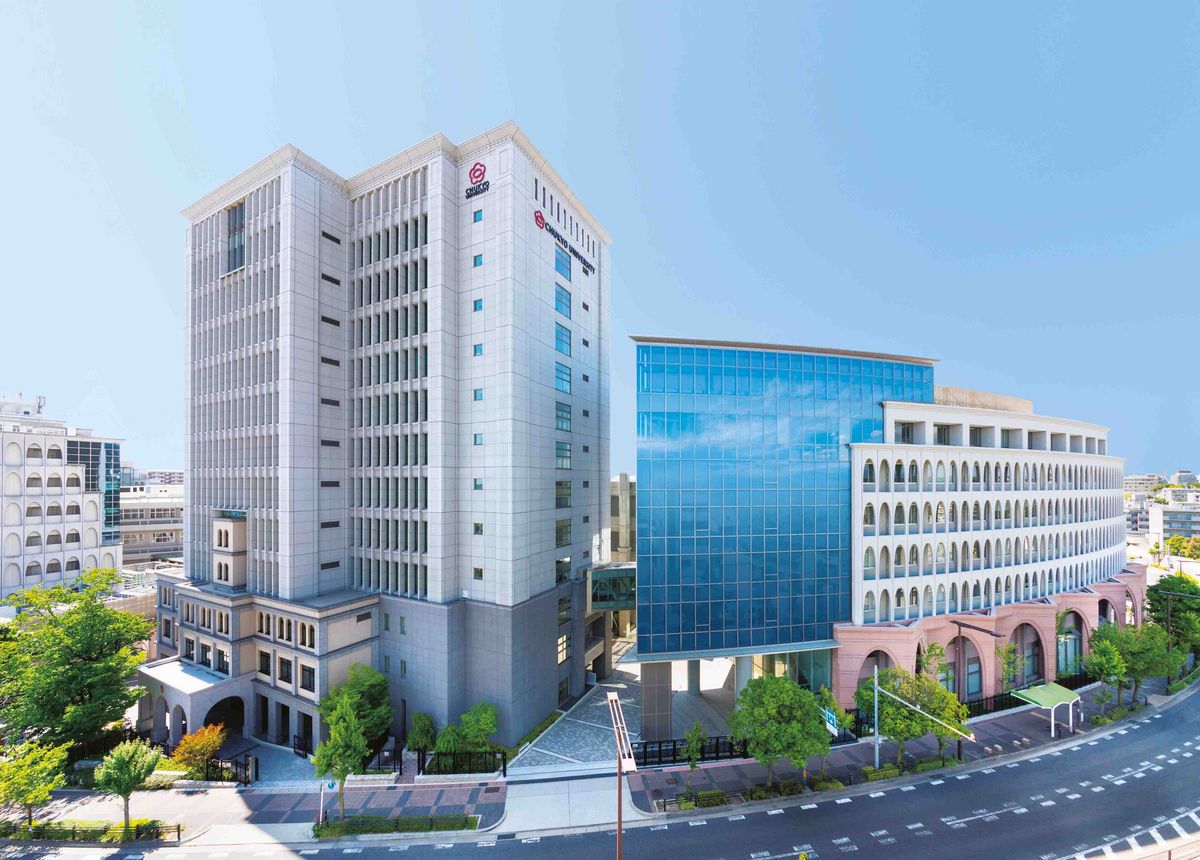 Ein hohes Gebäude auf dem Nagoya Campus der Chukyo Universität mit einer Fassade aus Glas und Stein. Das Gebäude hat eine rechteckige Form und ist mit dem Logo der Universität versehen. Bäume und eine Straße befinden sich im Vordergrund.