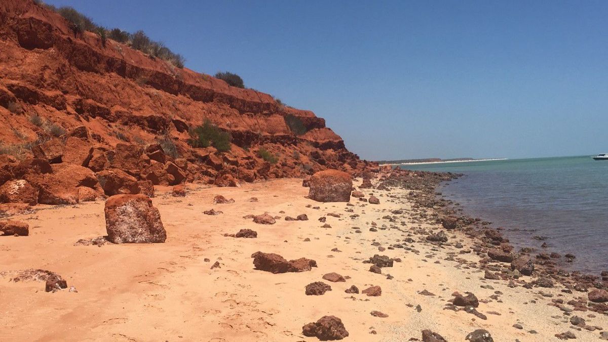 Das Bild zeigt eine Küstenlandschaft mit einem roten Felsenhang, der sich über einen Sandstrand erstreckt. Am Strand liegen große Felsbrocken verstreut. Das Wasser ist türkisblau und ruhig. Der Himmel ist klar und blau. Die Szene vermittelt ein Gefühl von Ruhe und Abgeschiedenheit.