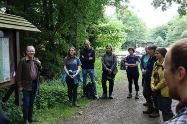 Das Foto zeigt eine Gruppe an Personen, die einer erzählenden Person gespannt zuhören. Sie befinden sich auf einer Exkursion im Gerdshorst vor einer Informationstafel im Wald.