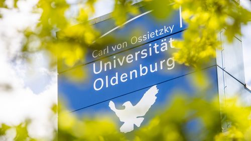 The photo shows the blue tower of building A1 to A4 with the logo of the University and the dove of peace; the leaves of a tree can be seen blurred in the foreground.