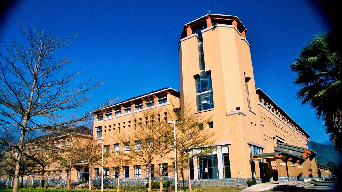 Das Bild zeigt ein Gebäude der National Dong Hwa University (NDHU). Das Gebäude hat eine helle, ockerfarbene Fassade mit vertikalen Akzenten und großen Fenstern. Im Hintergrund sind Berge und Bäume zu sehen, die dem Bild eine natürliche Kulisse verleihen. Die Szene wird durch einige Bäume im Vordergrund ergänzt, die dem Bild Tiefe verleihen.