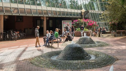 Auf dem Innenhof sitzen an Tischen und Bänken einige PErsonen locker herum, im Vordergrund die Brunnen, im Hintergrund die Glasfassade.