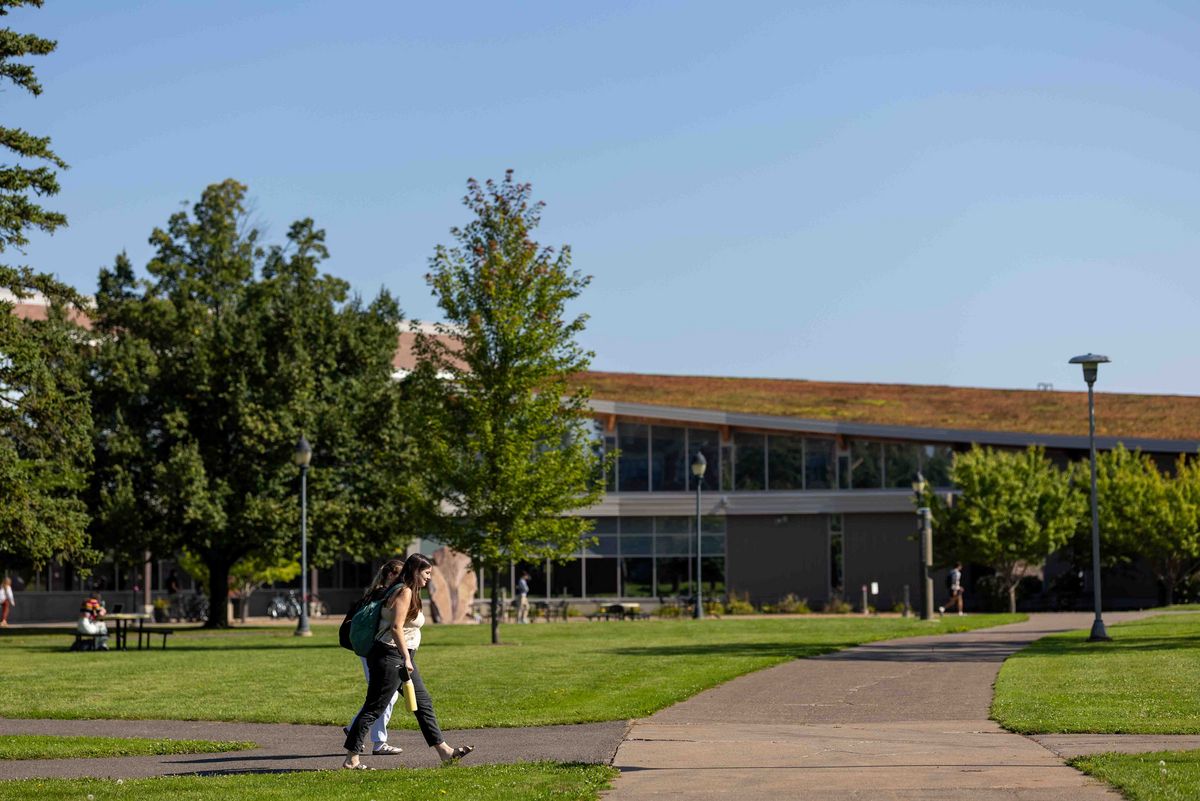 Campus  Zwei Studierende gehen auf einem Weg über den Campus. Bäume und ein Gebäude im Hintergrund.