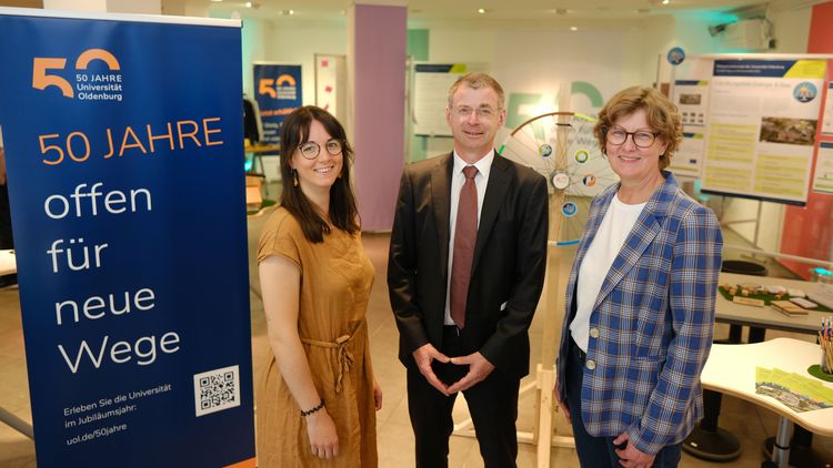 Das Bild zeigt den Store von innen. Links steht ein Roll-Up mit dem Schriftzug "50 Jahre offen für neue Wege" und dem Jubiläumslogo der Uni Oldenburg. Rechts daneben stehen Anna Krämer, Ralf Grüttemeier und Corinna Dahm-Brey und lächeln in die Kamera. Im Hintergrund sind Poster und Stellwände mit Informationen zu erkennen.