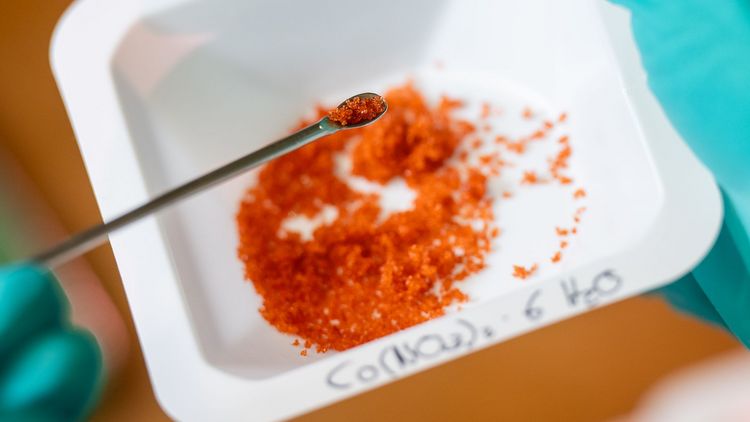Close-up of a bowl of rust-coloured metallic salt. In the foreground is a spatula with a small amount of salt on it. 