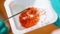 Close-up of a bowl of rust-coloured metallic salt. In the foreground is a spatula with a small amount of salt on it. 