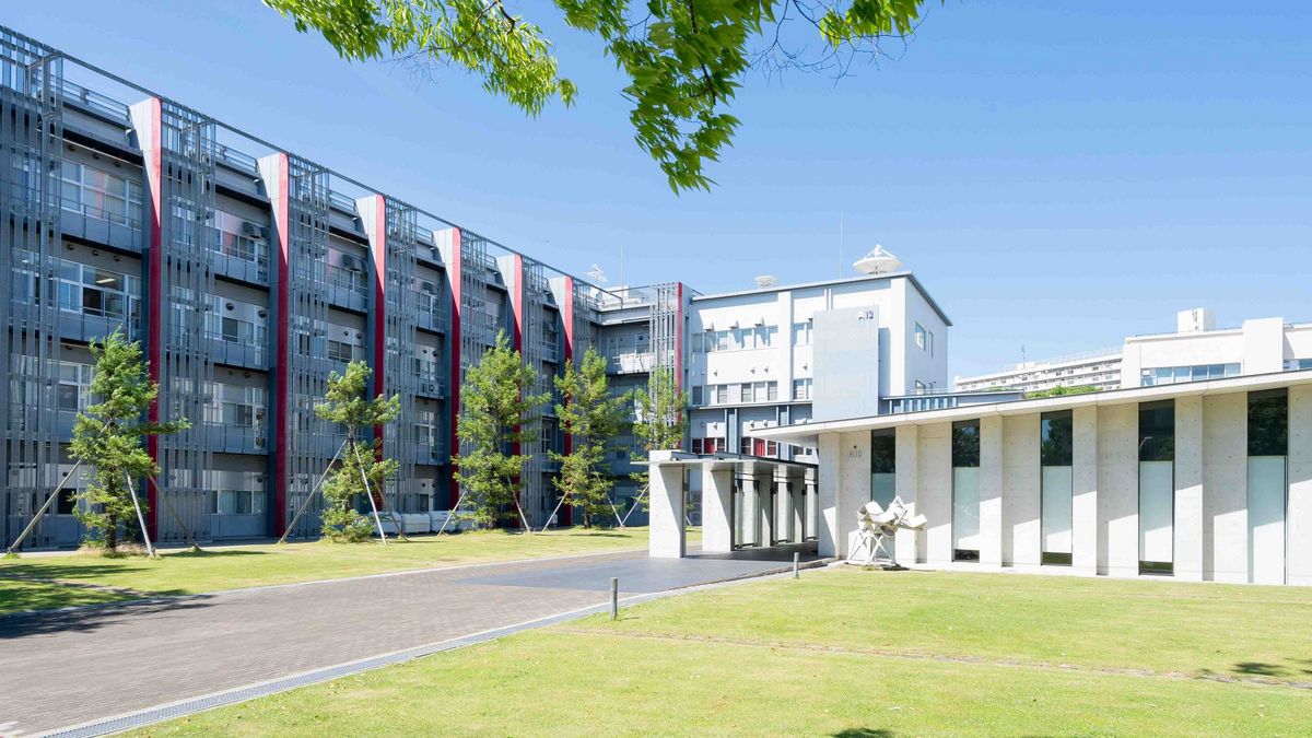 Das Bild zeigt einen modernen, weitläufigen Gebäudekomplex der Osaka Metropolitan University, der durch eine Kombination aus grauen und roten Elementen gekennzeichnet ist. Der Gebäudekomplex besteht aus mehreren miteinander verbundenen Strukturen mit einer Fassade aus vielen kleinen Fenstern und Balkonen. Vor dem Gebäude erstreckt sich eine große Grünfläche mit einem gewundenen Weg und einigen Bäumen, die eine offene und einladende Atmosphäre schaffen