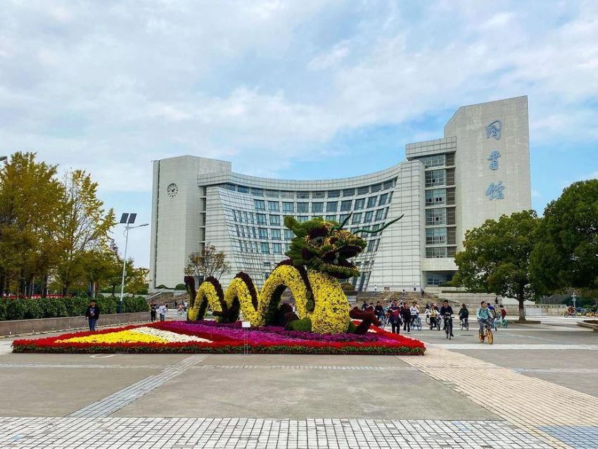 Das Bild zeigt ein großes, modernes Gebäude der Shanghai University an einem sonnigen Tag. Vor dem Gebäude befindet sich ein farbenfrohes Blumenbeet in Form eines Drachen, umgeben von Bäumen und einer gepflasterten Fläche. Mehrere Menschen sind zu sehen, die sich auf dem Campus bewegen, einige stehen vor dem Gebäude und andere gehen oder fahren Fahrrad vorbei.