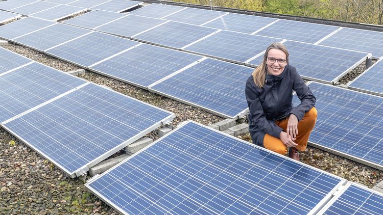 Eine Frau hockt auf einem Flachdach, das mit Solarmodulen bedeckt ist und lächelt in die Kamera.